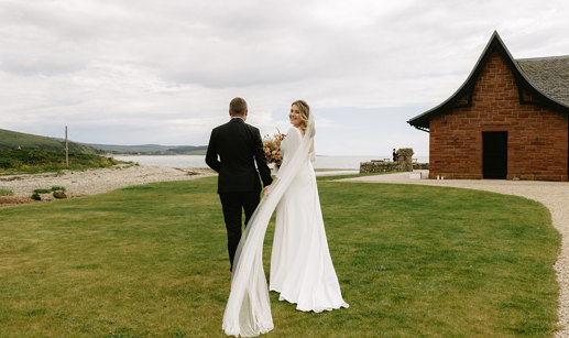 A bride and groom stroll hand in hand on lush grass towards the sea with a red brick building with pitched roof on right