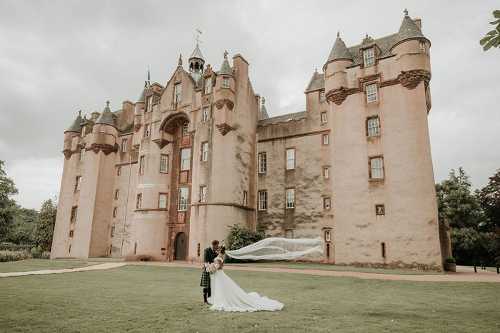 A groom in a kilt and a bride in a dress with a train stand in front of a large castle 