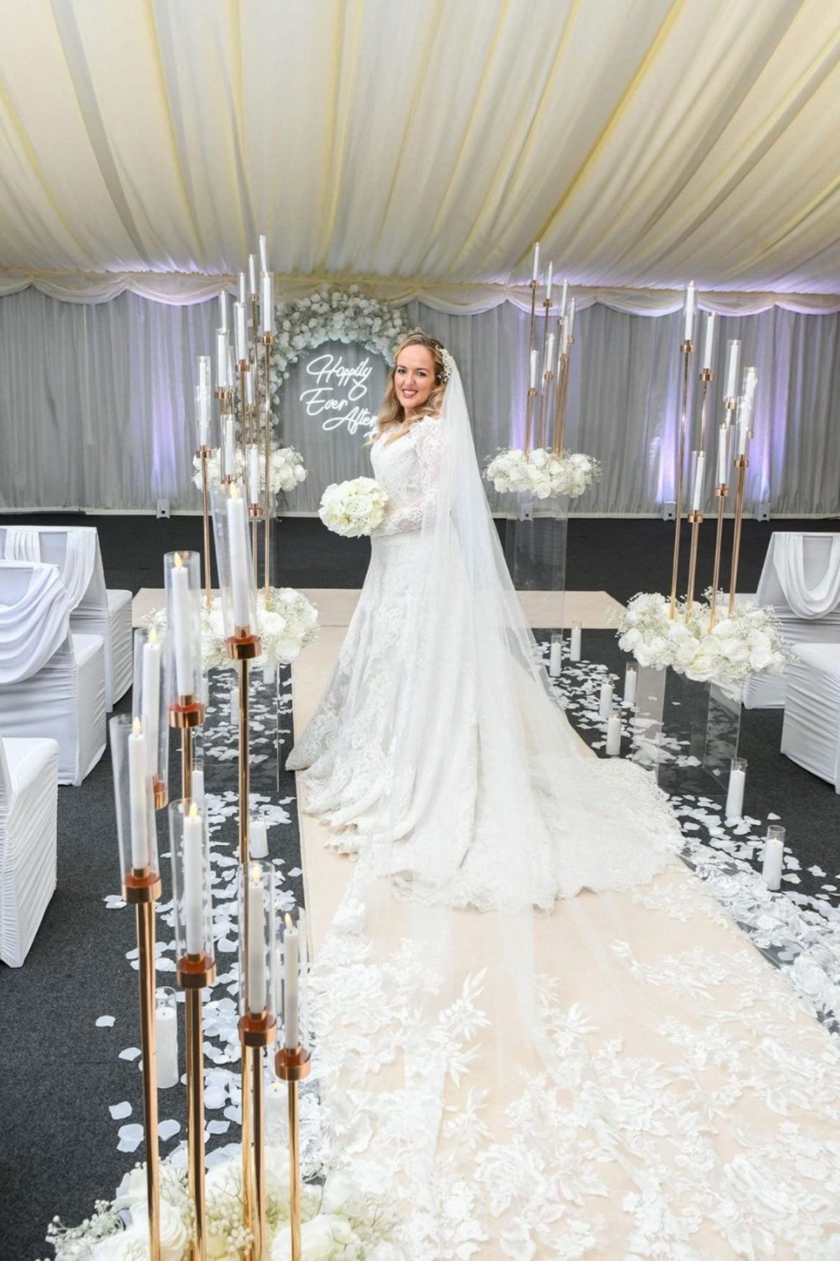 Bride in a lace wedding dress standing beneath a white draped aisle setup with candles and floral décor at Geilsland Estate.