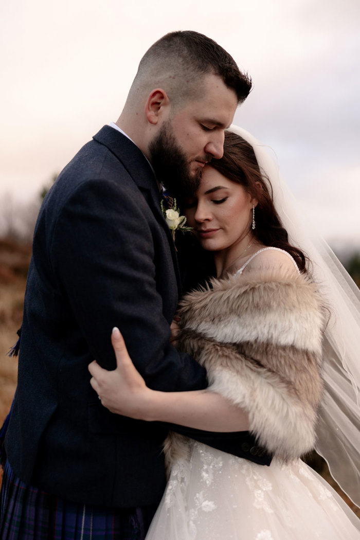 a bride wearing a faux fur wrap, veil and ivory dress snuggles into a groom wearing a blue kilt and tweed jacket. They have their eyes closed and the light is soft