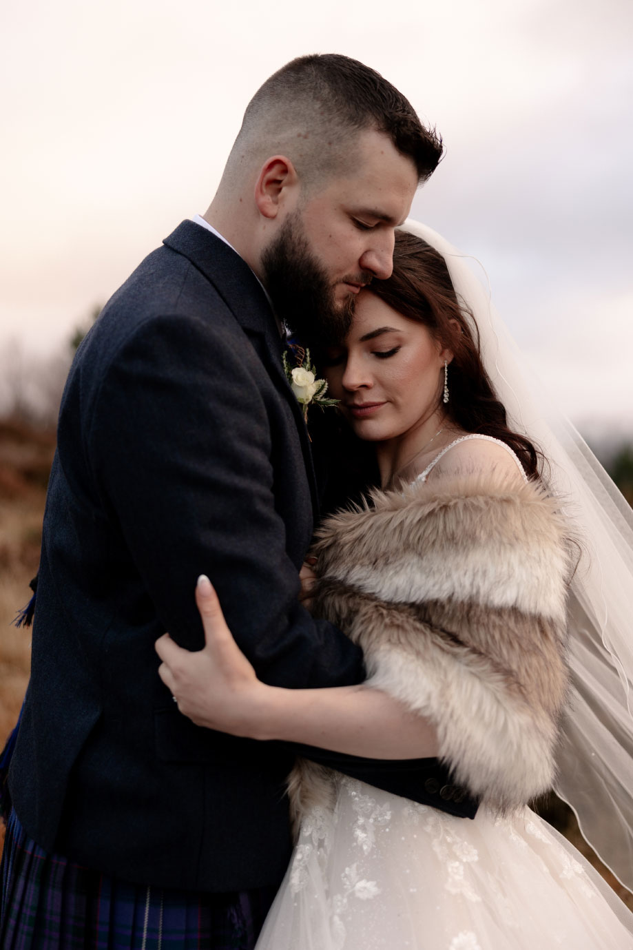 a bride wearing a faux fur wrap, veil and ivory dress snuggles into a groom wearing a blue kilt and tweed jacket. They have their eyes closed and the light is soft