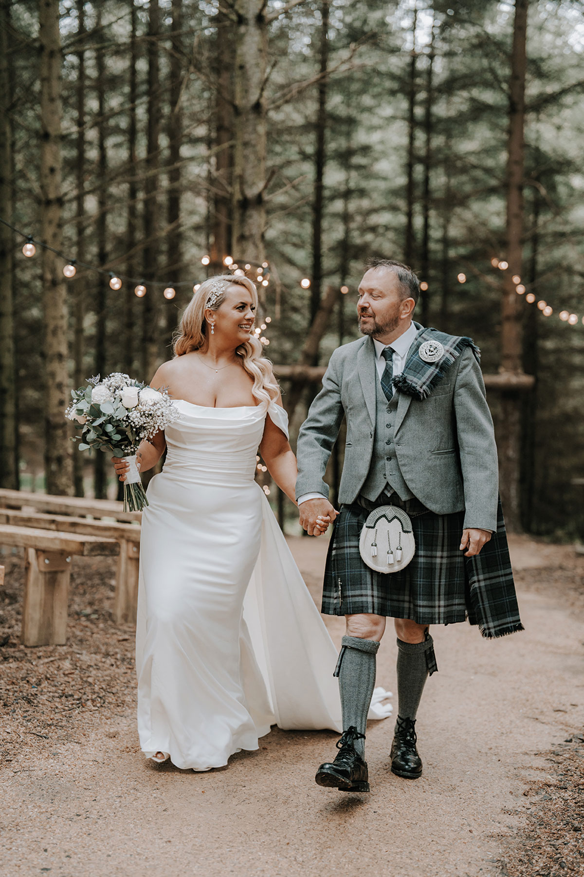 Bride and groom holding hands while walking along a woodland path decorated with lights.