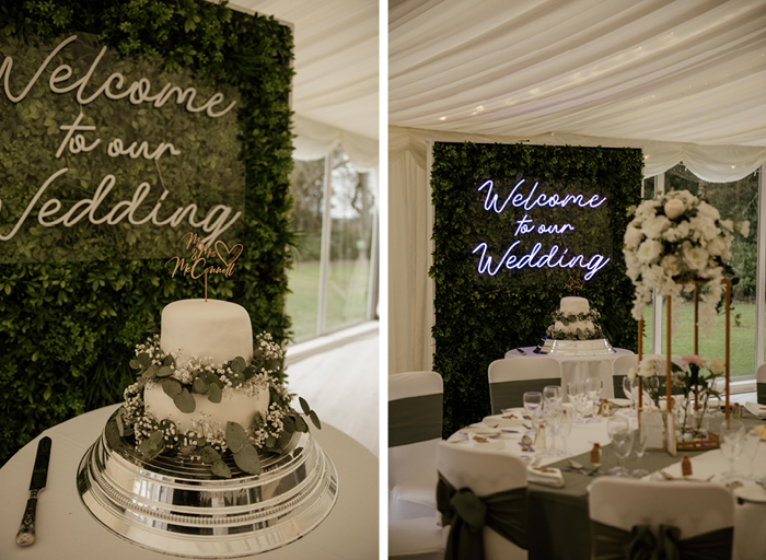 A white two-tier cake surrounded by foliage and white flowers sitting in front of a neon 'welcome to our wedding' sign