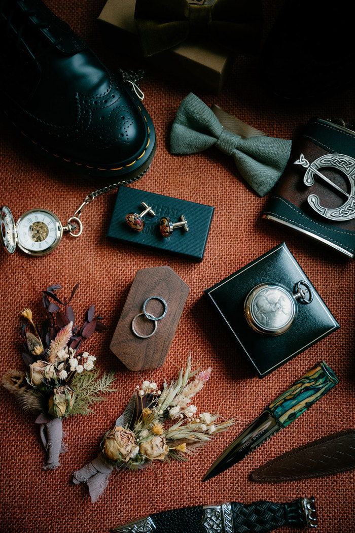 black brogue shoes, a green bow tie, wedding rings, sgian dubhs and flower buttonholes sitting on orange woven fabric