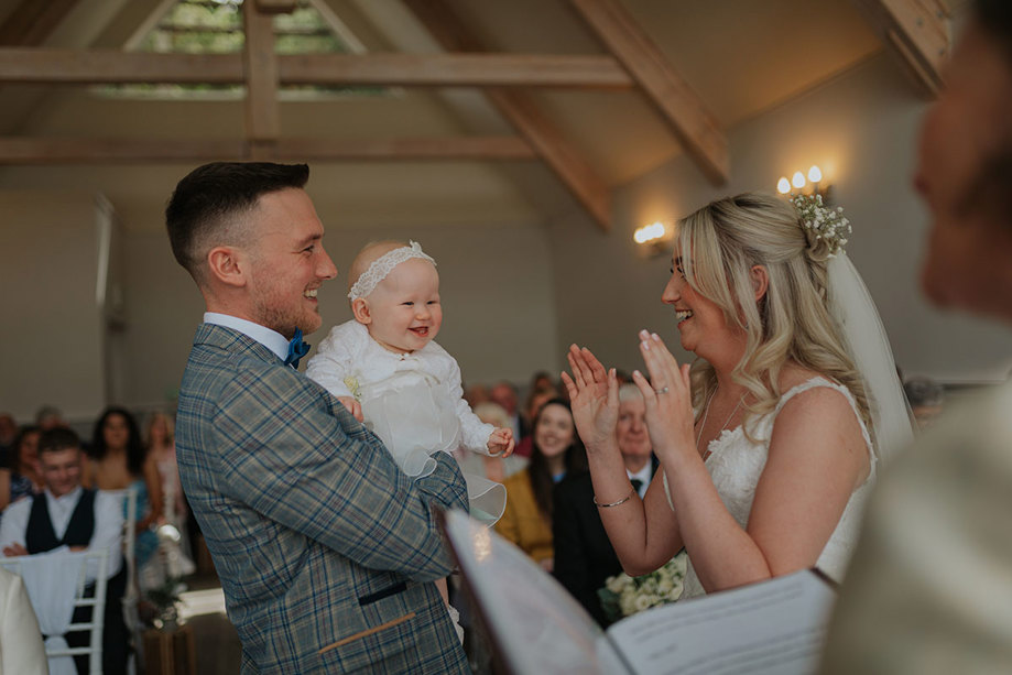A Bride Claps Her Hands While Groom Smiles And Holds Young Child