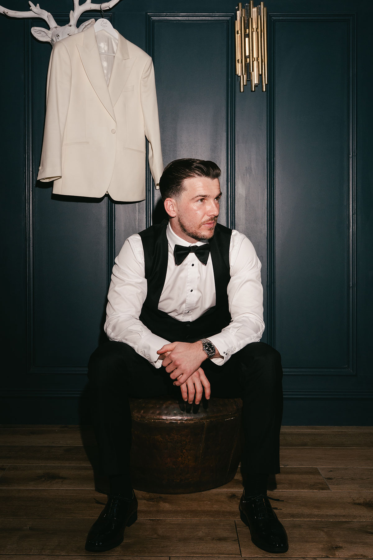 Groom portrait seated in tuxedo with white jacket hanging behind at Enterkine Country House wedding