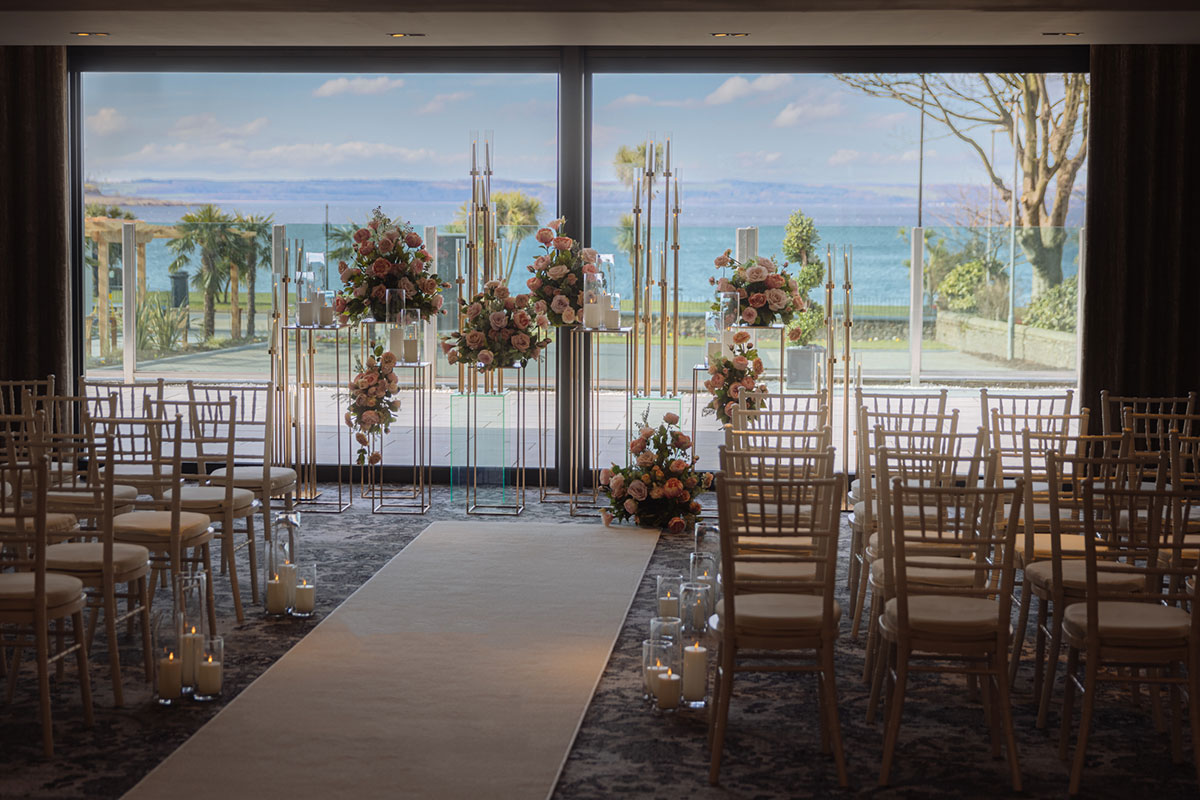 cream aisle runner leads up to front of room with different levels of heightened floral displays in front of window wall looking out to an ocean view