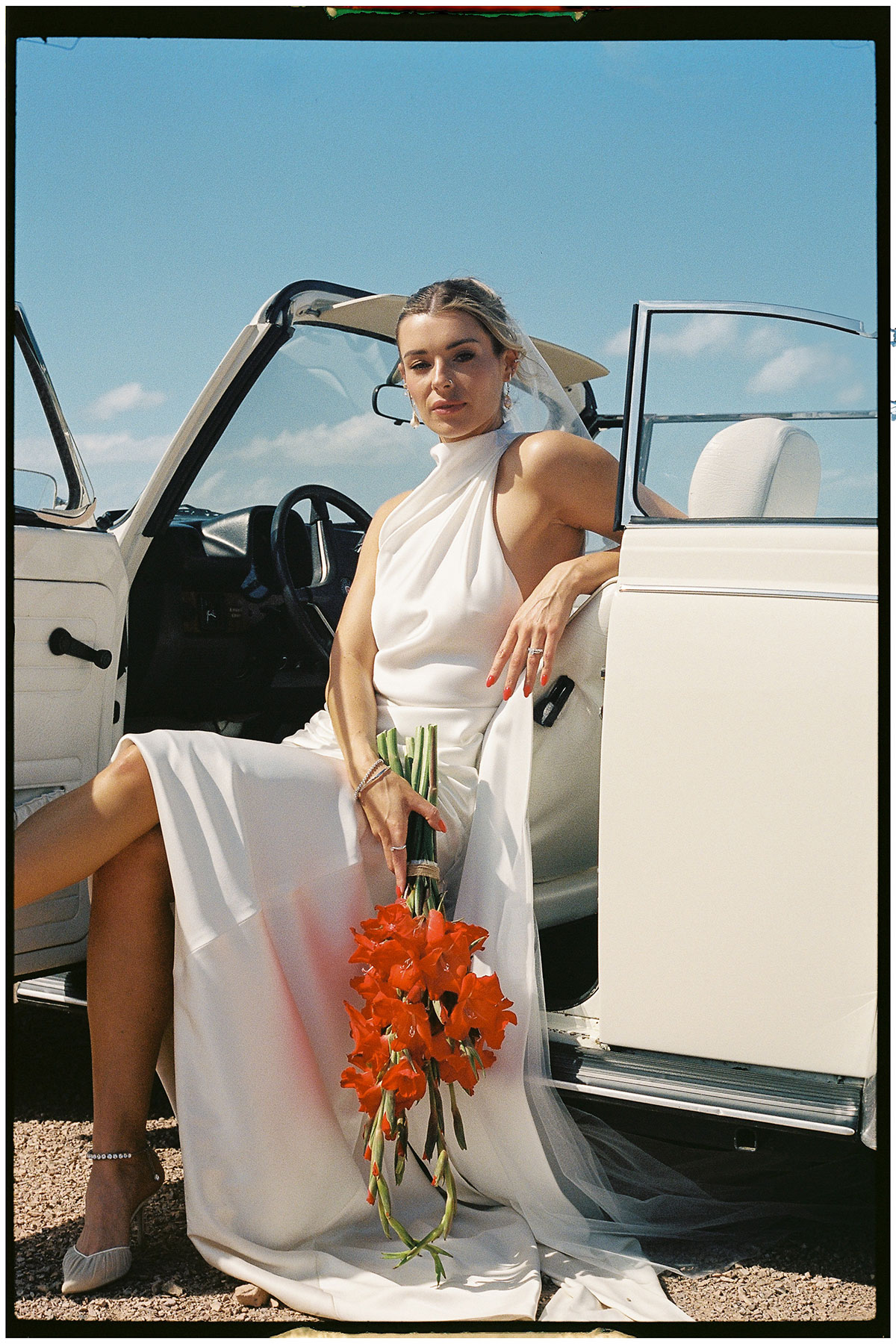 Bride poses in a vintage white Volkswagen Beetle convertible holding a bright red bouquet at Kinkell Byre wedding venue in Fife.