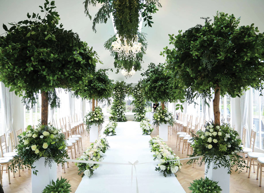 a wedding set up in the conservatory at Carlowrie Castle by Jordana Events and Weddings with white aisle runner, trees in pots either side of the aisle, green and white flower arrangements on floor and green arrangements hanging from the ceiling