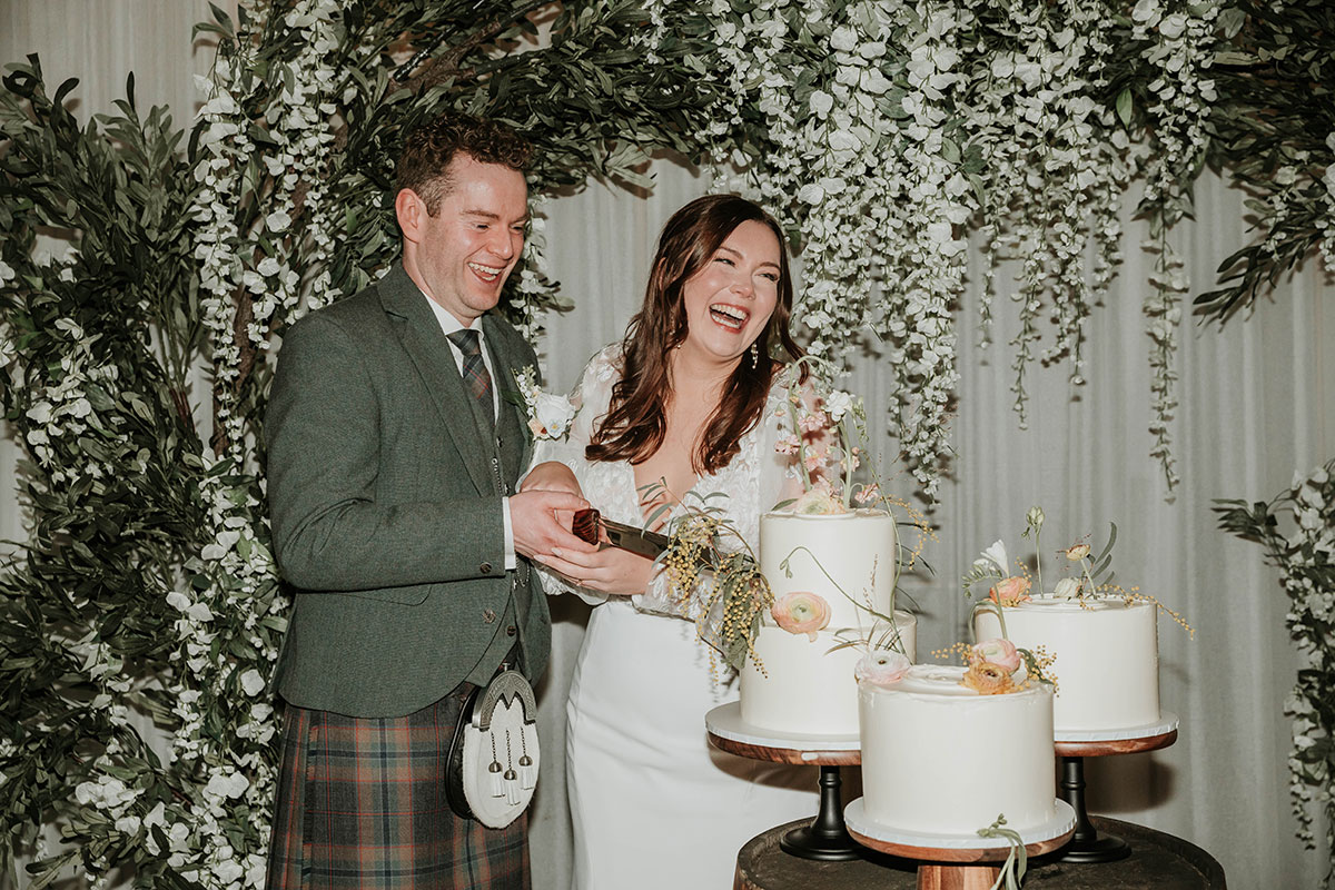Bride and groom laughing while cutting wedding cake decorated with flowers