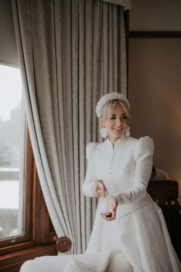 A bride wearing a textured dress with a matching high neck jacket applying perfume 