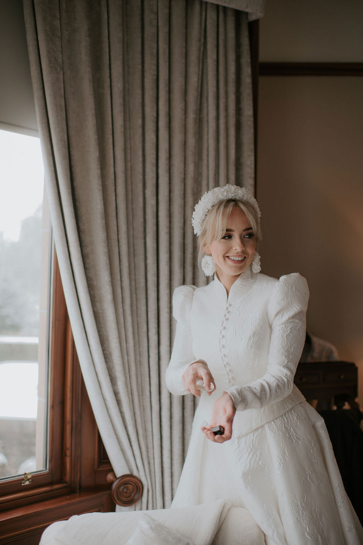 A bride wearing a textured dress with a matching high neck jacket applying perfume
