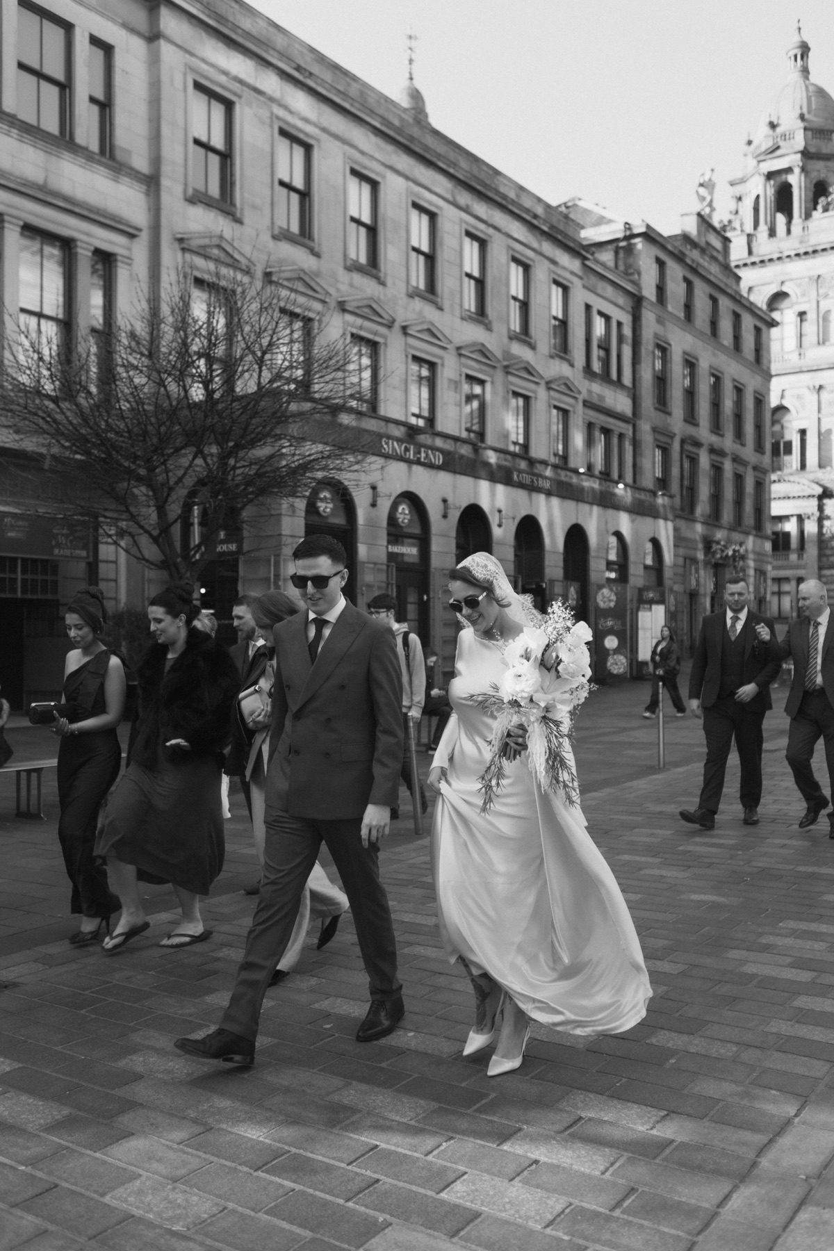 Newlyweds walking through Glasgow city centre with their wedding party following behind.