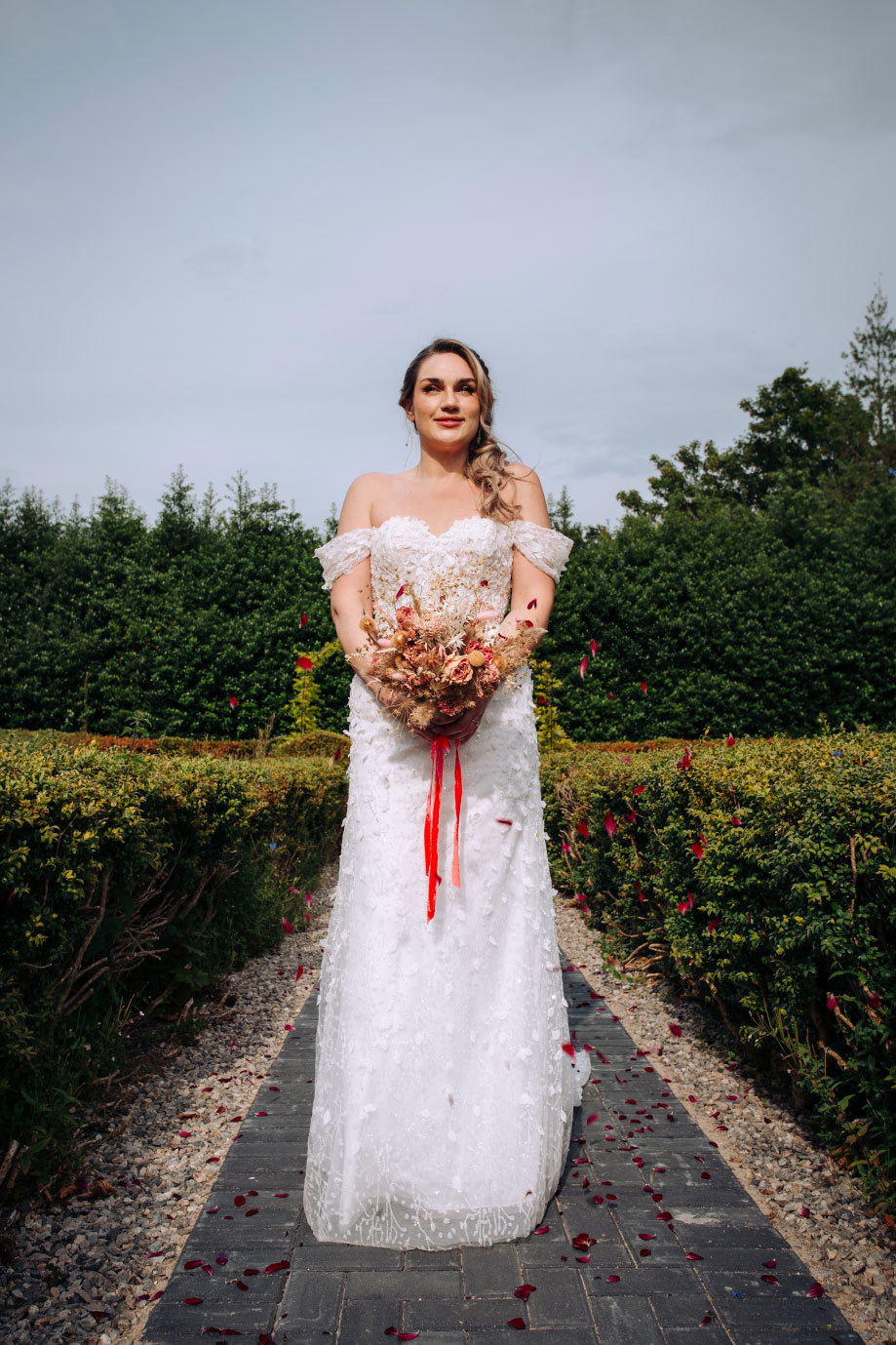 bride stands in an outdoor aisle wearing an off the shoulder gown and holding a dried floral bouquet at the centre of her waist