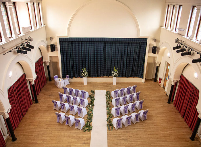 A hall with wooden floors, white walls, white chairs facing a stage which has a green and blue tartan curtain 