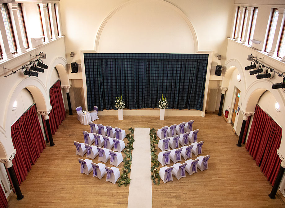 A hall with wooden floors, white walls, white chairs facing a stage which has a green and blue tartan curtain