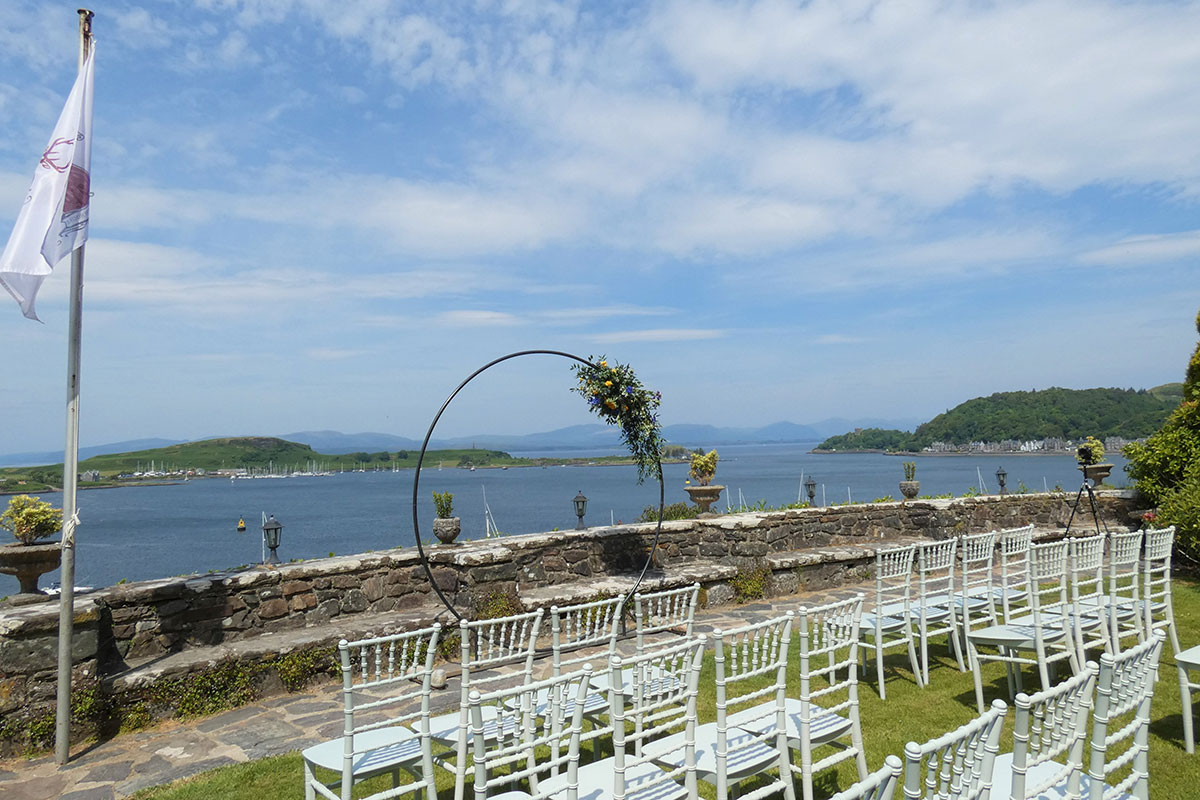 An outdoor ceremony setup with white chairs facing panoramic sea views and a circular floral arch.