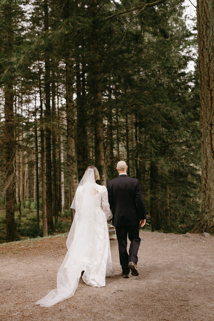 bride in white floral wedding dress with trailing veil and groom in black suit, walk hand in hand, both with their backs to the camera, towards a forrest of tall slim trees