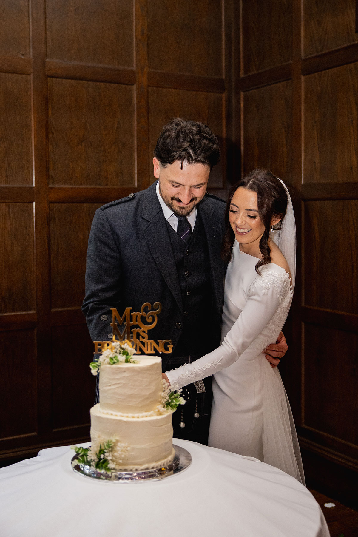 Couple smile as they cut their two-tier white wedding cake with a “Mr & Mrs Browning” topper in a wood-panelled room