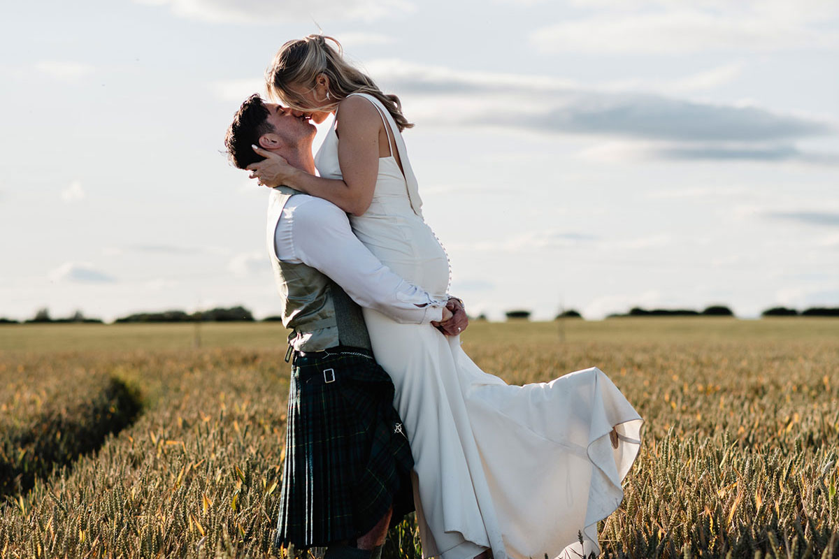 Groom in a kilt lifts the bride in a white dress as they kiss