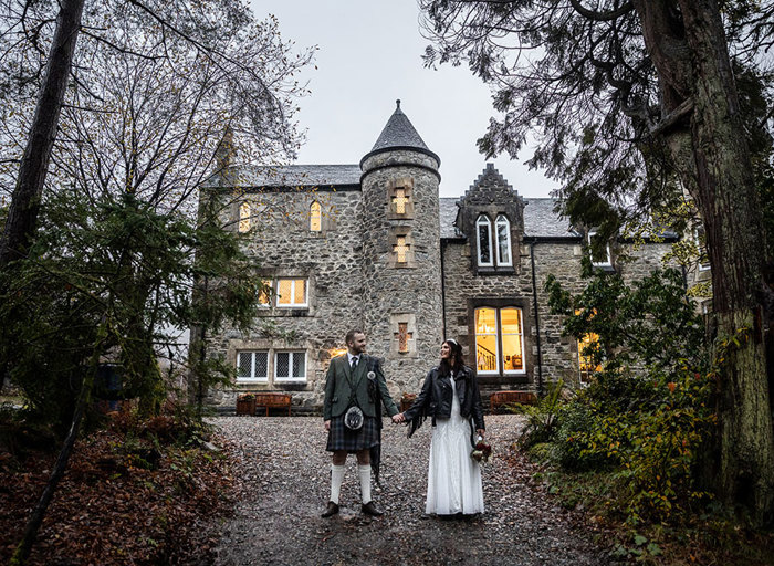 a Scottish wedding day couple holding hands outside a small castle-like building