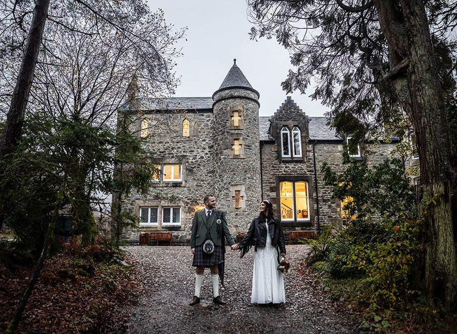a Scottish wedding day couple holding hands outside a small castle-like building
