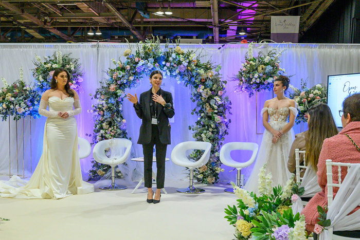 a person making a presentation in front of a floral arch with two brides standing either side