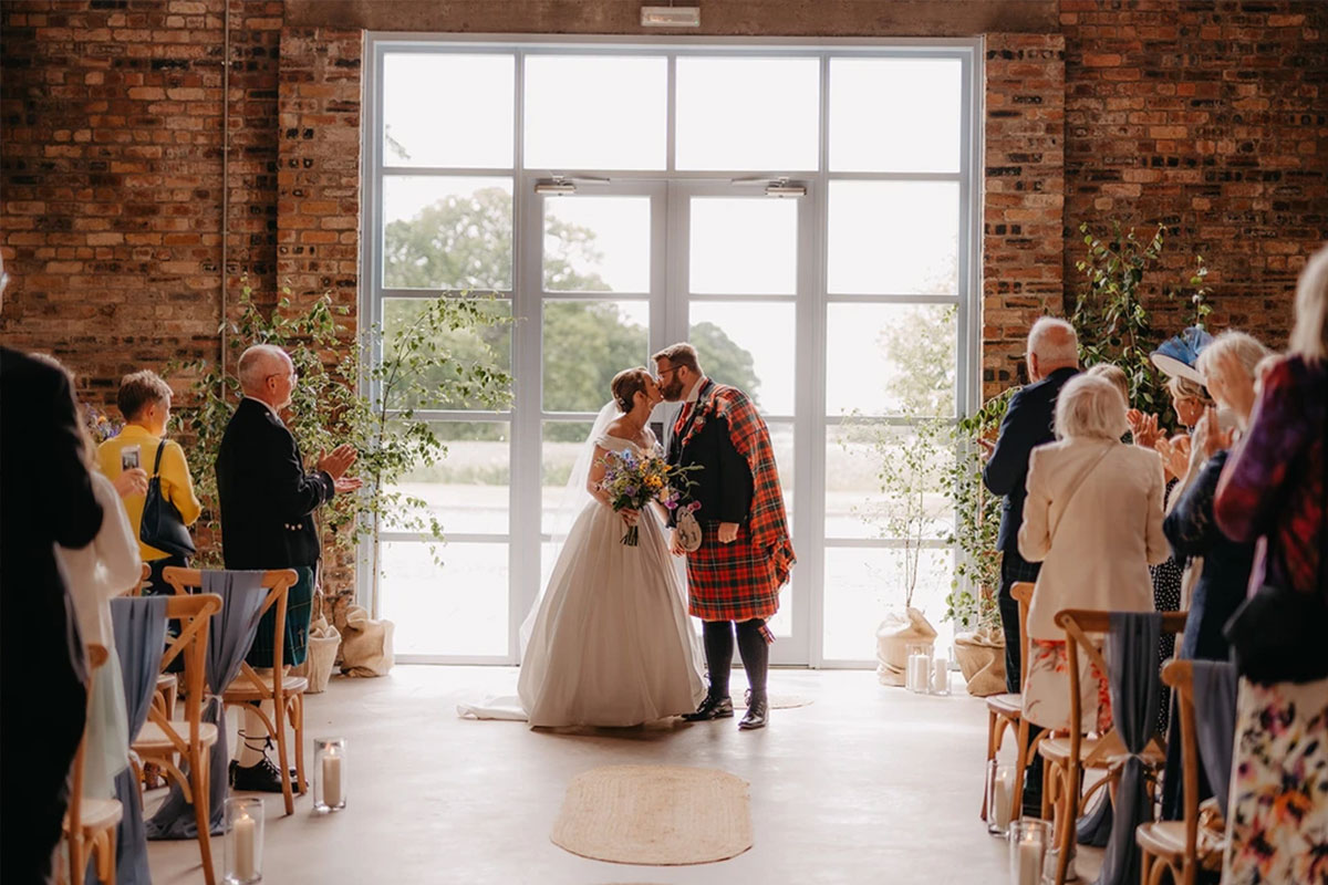 A bride and groom kiss during their ceremony set against glass doors and a brickwork wallas their guests stand and clap