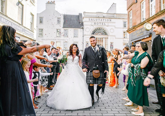 A bride in a ballgown wedding dress and a man in a kilt walking down a street as the wedding guests throw confetti over them