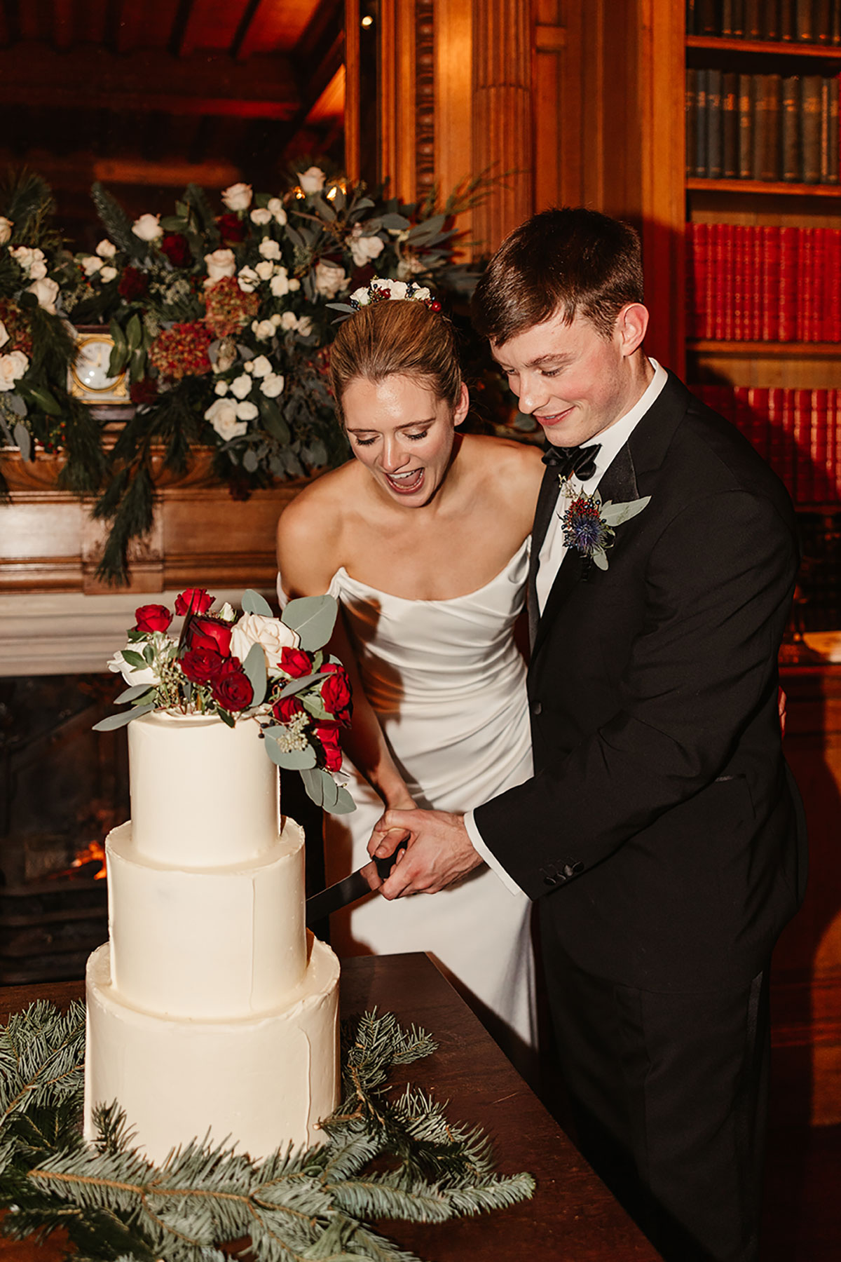 Bride and groom cutting a three-tier ivory wedding cake decorated with red and white flowers and winter greenery.