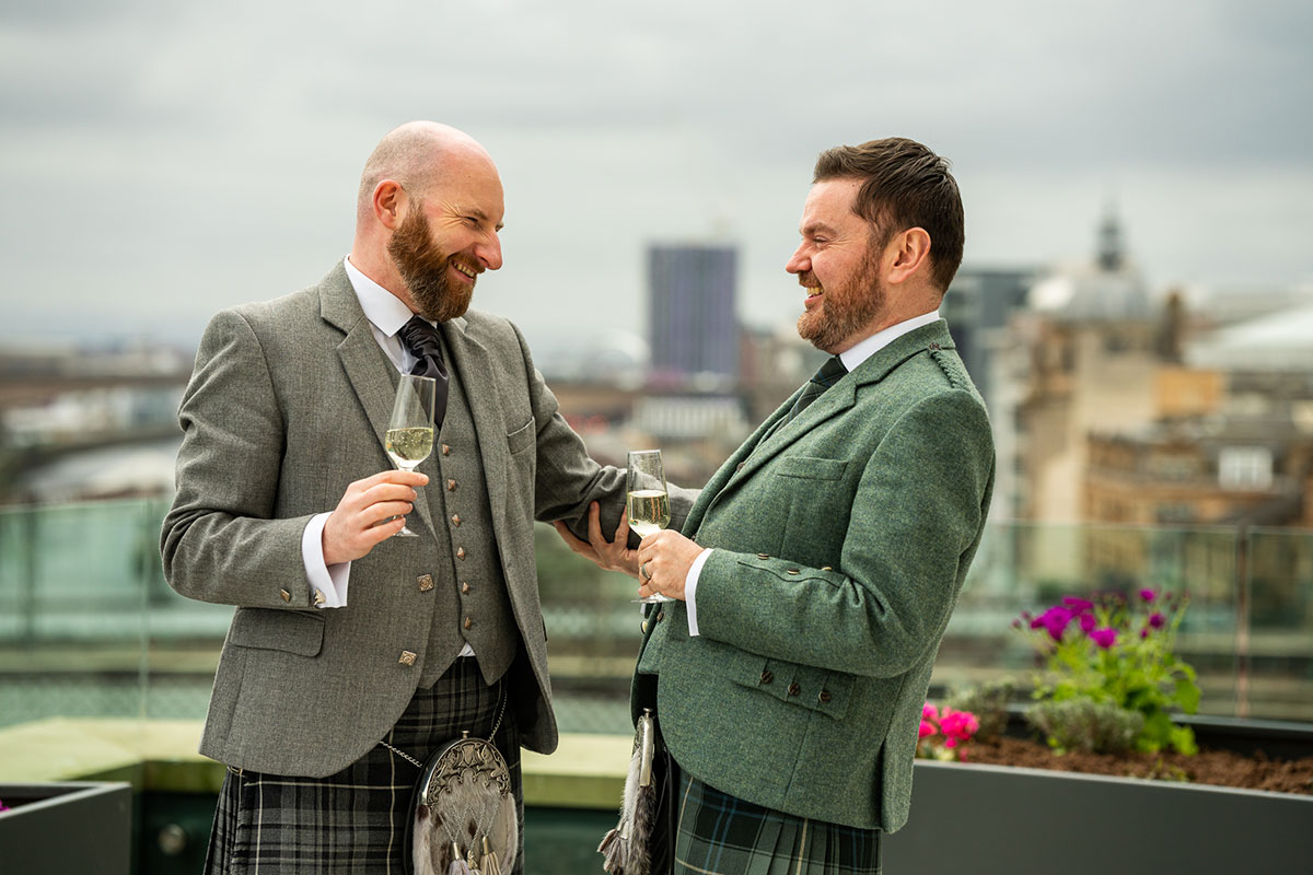 two grooms smile at one another while wearing kilts and holding a glass of fizz at clayton hotel glasgow