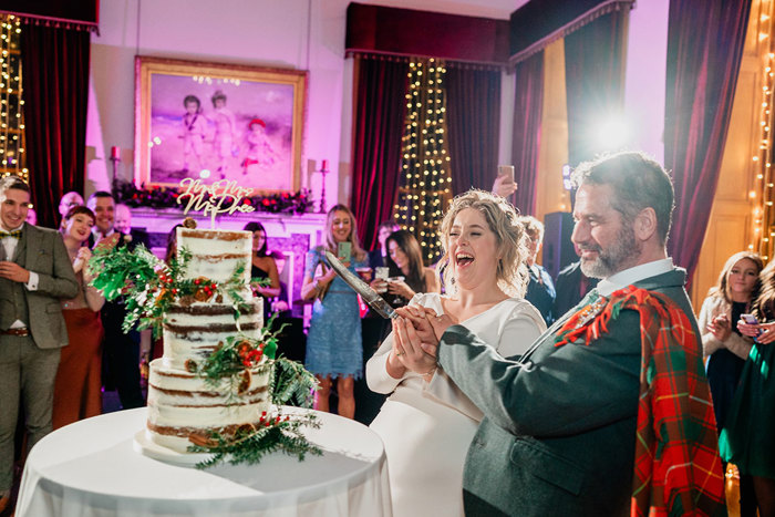 bride in white wedding dress and groom in red and green tartan outfit stand in front of three tier cake holding onto the same large cake knife