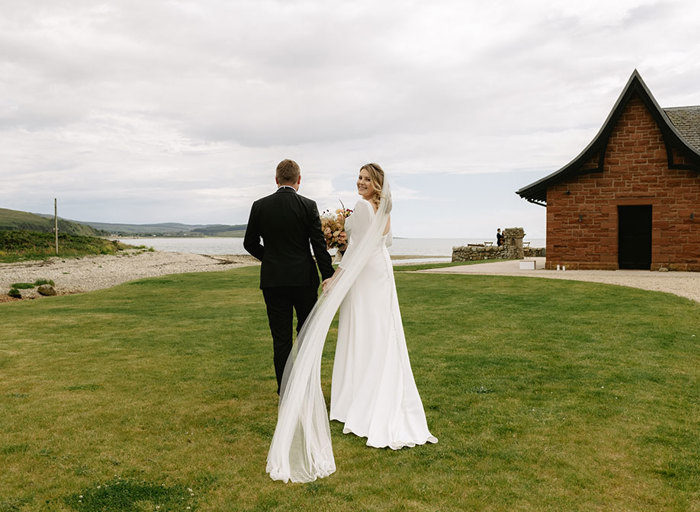 A bride and groom stroll hand in hand on lush grass towards the sea with a red brick building with pitched roof on right