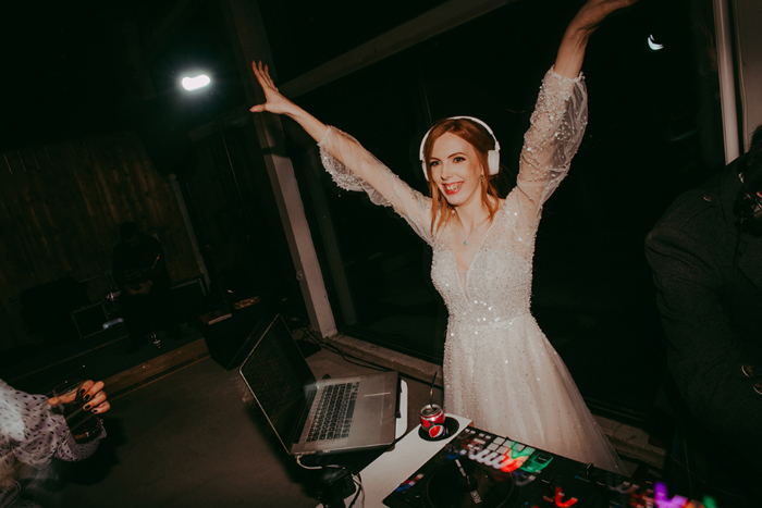 Bride throws her hands in the air behind the DJ decks wearing headphones 