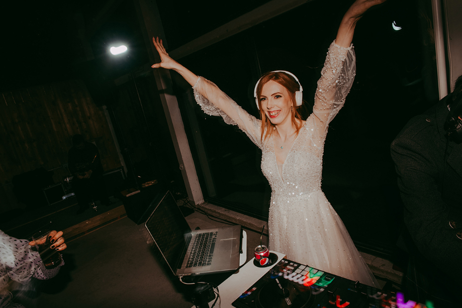 Bride throws her hands in the air behind the DJ decks wearing headphones