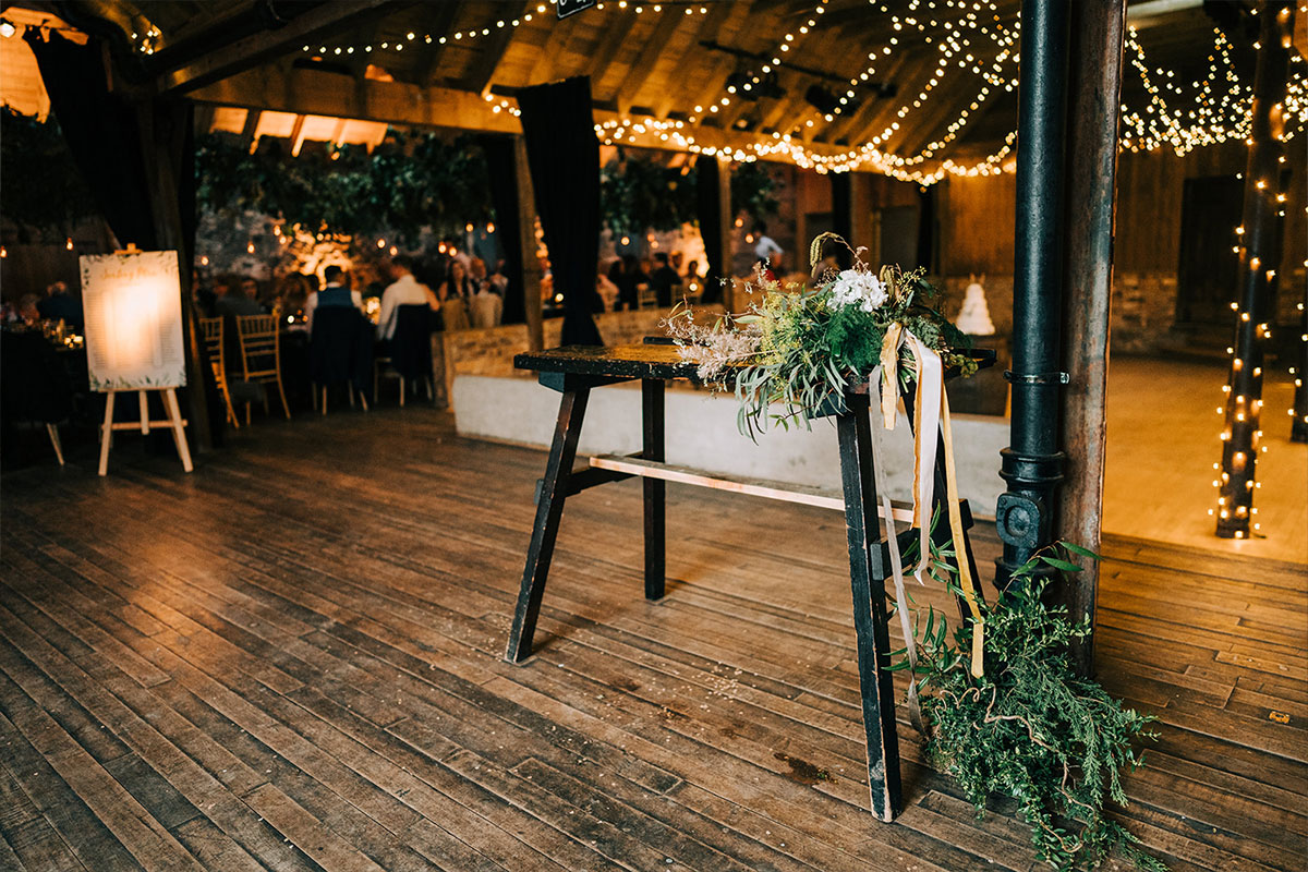 A barn space with fairylights handing from the roof beams in the background and a wooden table with greenery on it in the foreground