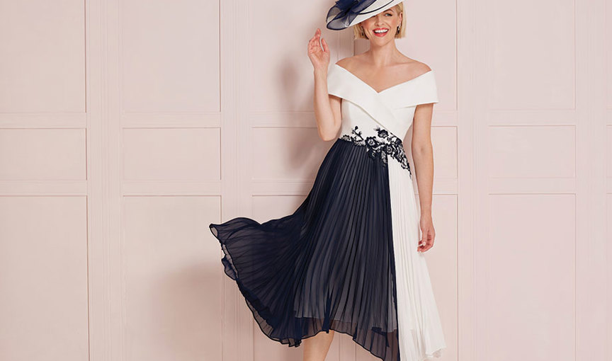 A person wearing an ivory and navy off the shoulder dress with pleated skirt standing against a pale pink panelled wall