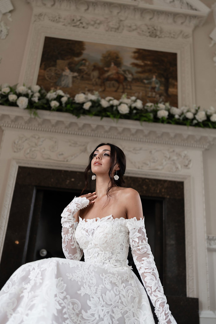 ground up view of woman in off the shoulder long sleeved white floral wedding dress crouching down in front of unused white marble fireplace decorated with white a shelf of foliage and white roses