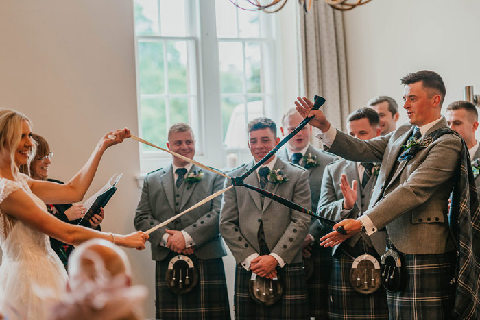 A bride and groom tying the knot with two pieces of fabric, one dark blue and one white while groomsmen in kilts look on