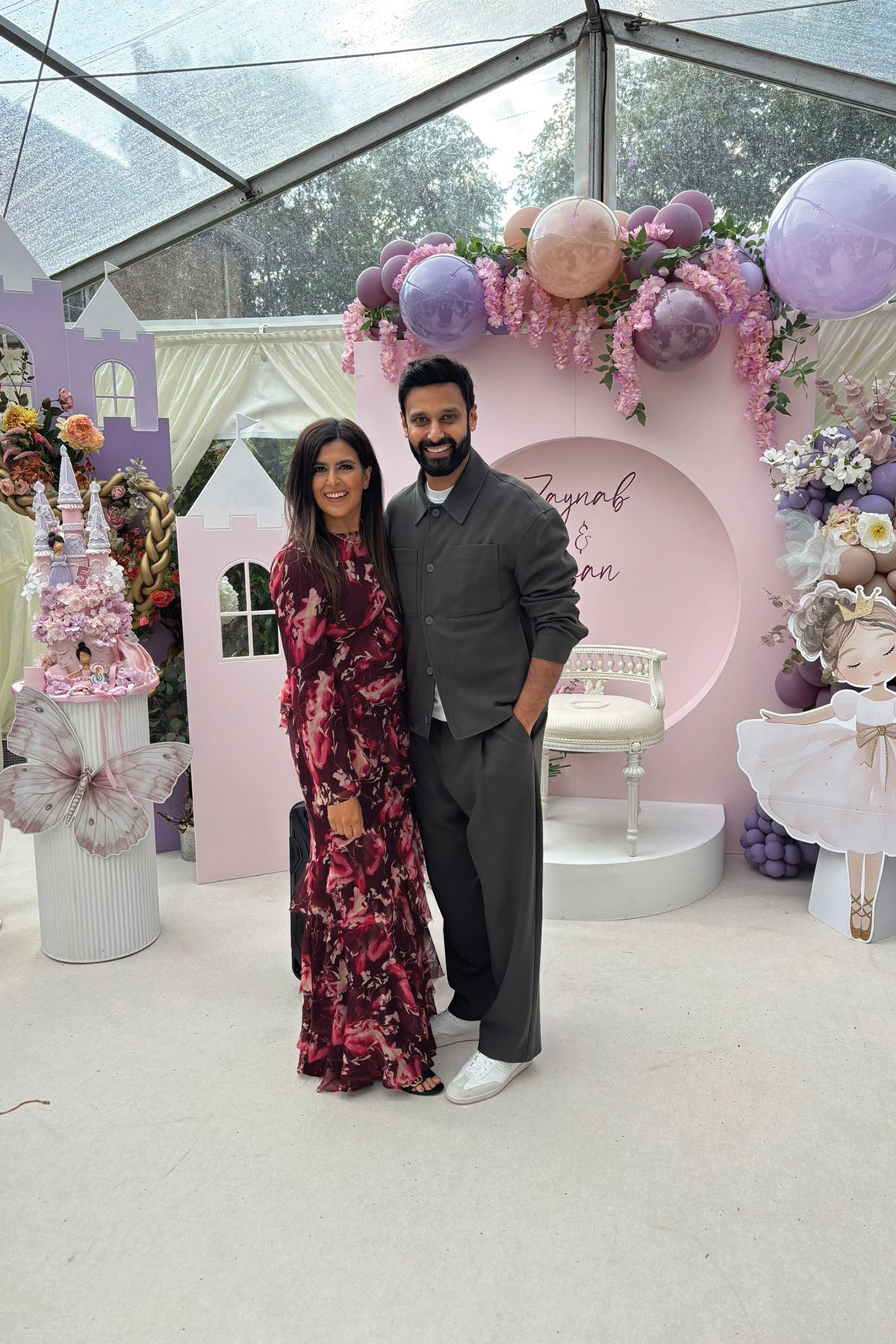 Couple standing in front of a pastel pink and lilac balloon display at a child’s birthday party, with castle-themed decorations and a personalised backdrop inside a marquee