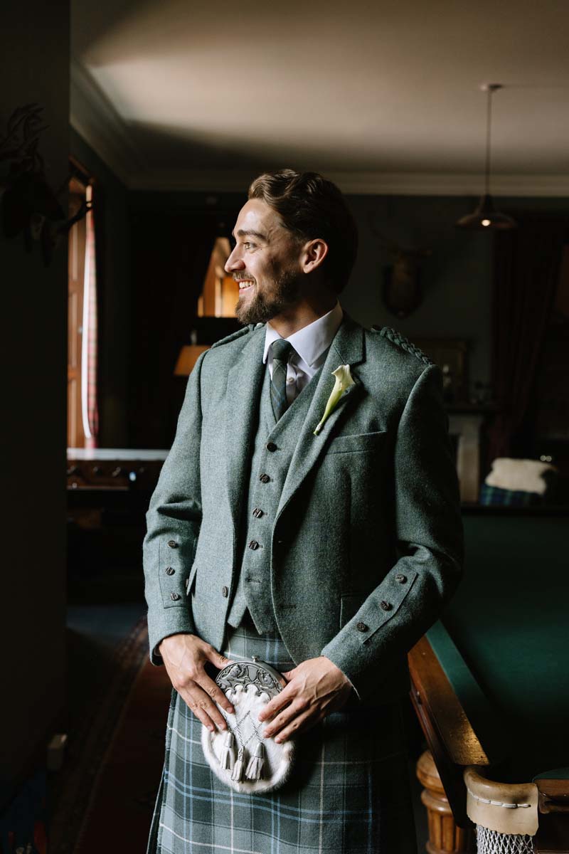 Groom Louis poses inside Cambo House wearing a green tweed jacket and traditional tartan kilt.