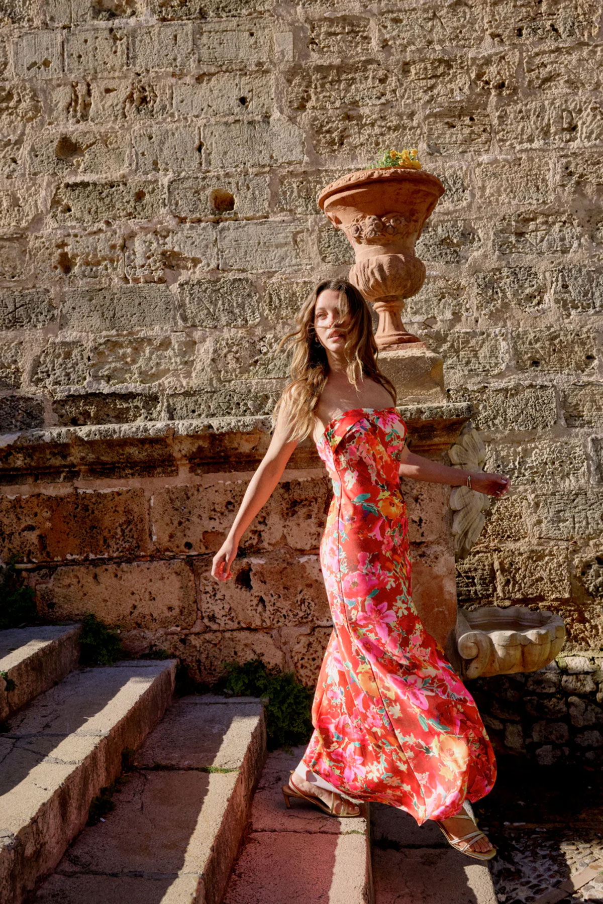 Red floral strapless maxi dress with flowing skirt worn outdoors on stone steps in warm sunlight
