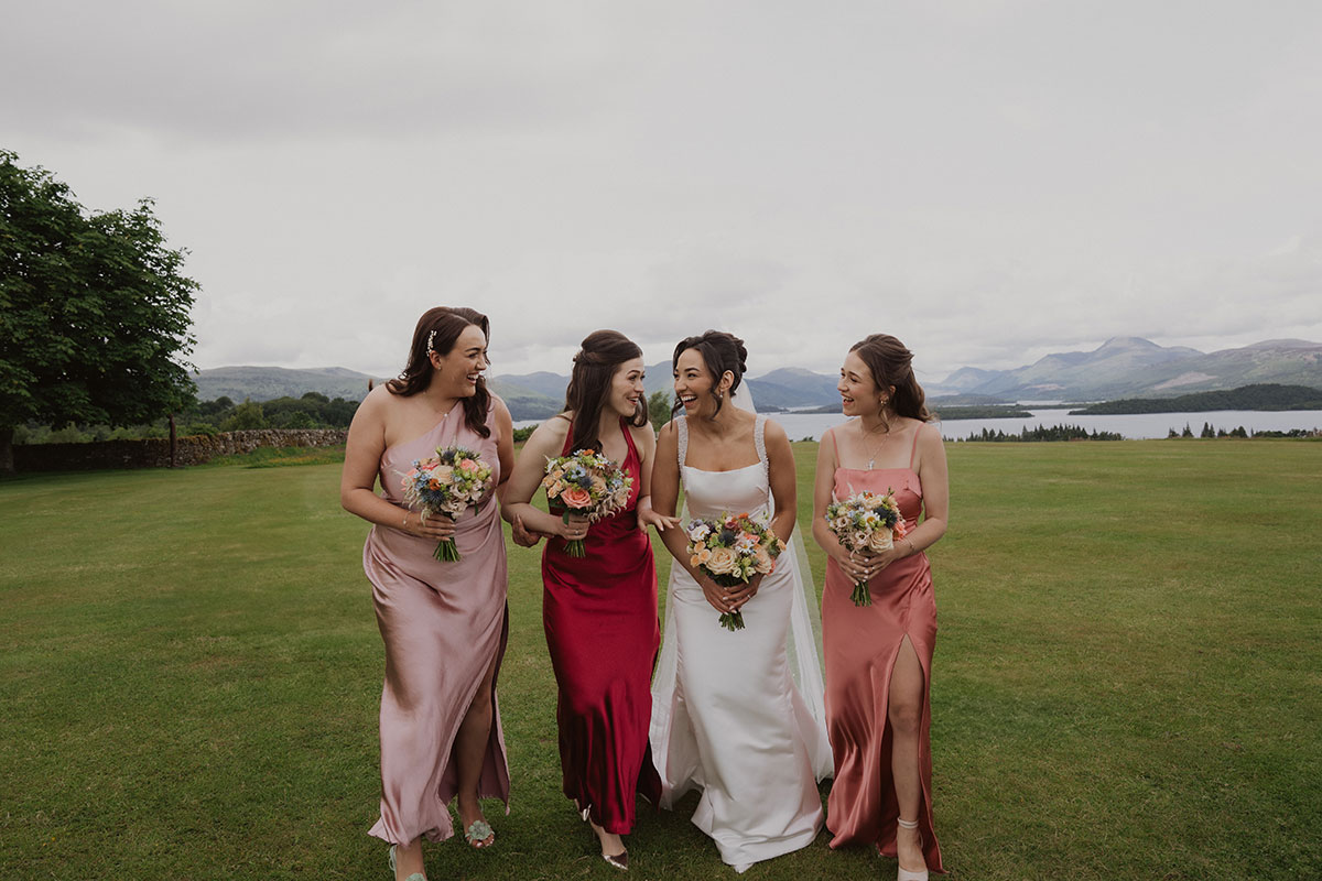 Bride walking with bridesmaids in satin pink and red dresses holding colourful bouquets overlooking Scottish loch