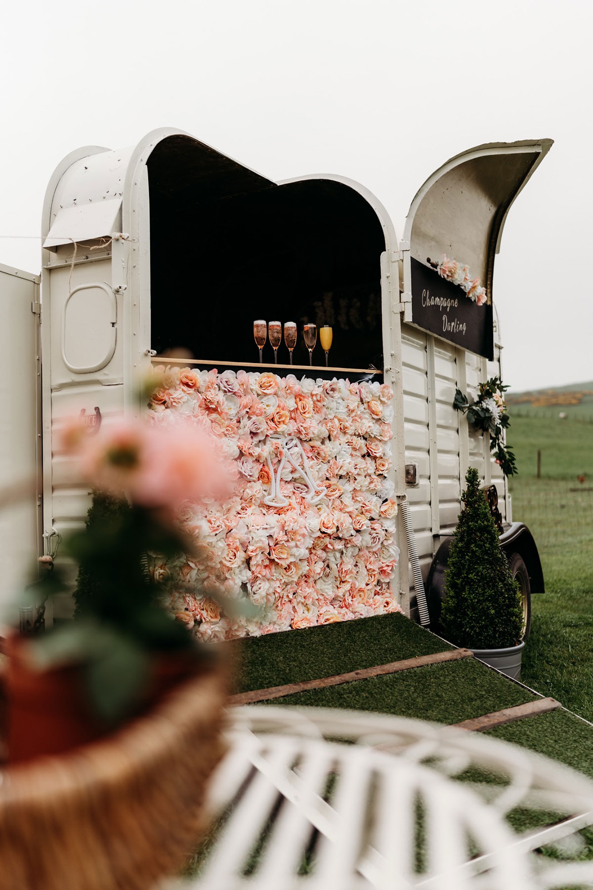 a grey white horsebox is decorated with a pink and white faux flower wall that has five glasses of fizz sat above it and a faux grass ramp below it