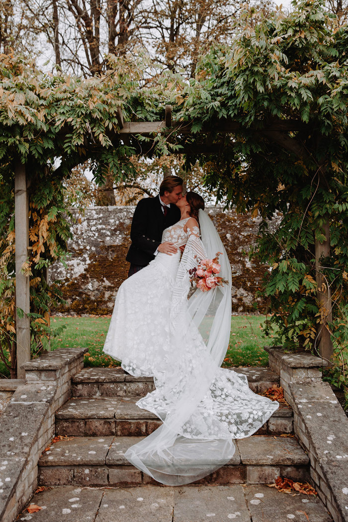 bride and groom dip kiss under a brown and green foliage covered square arch at the top of four stone tile steps