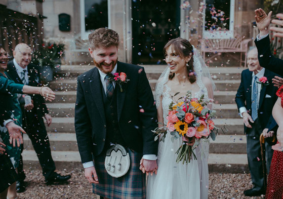 a bride and groom walking through a colourful confetti shower outside Netherbyres House