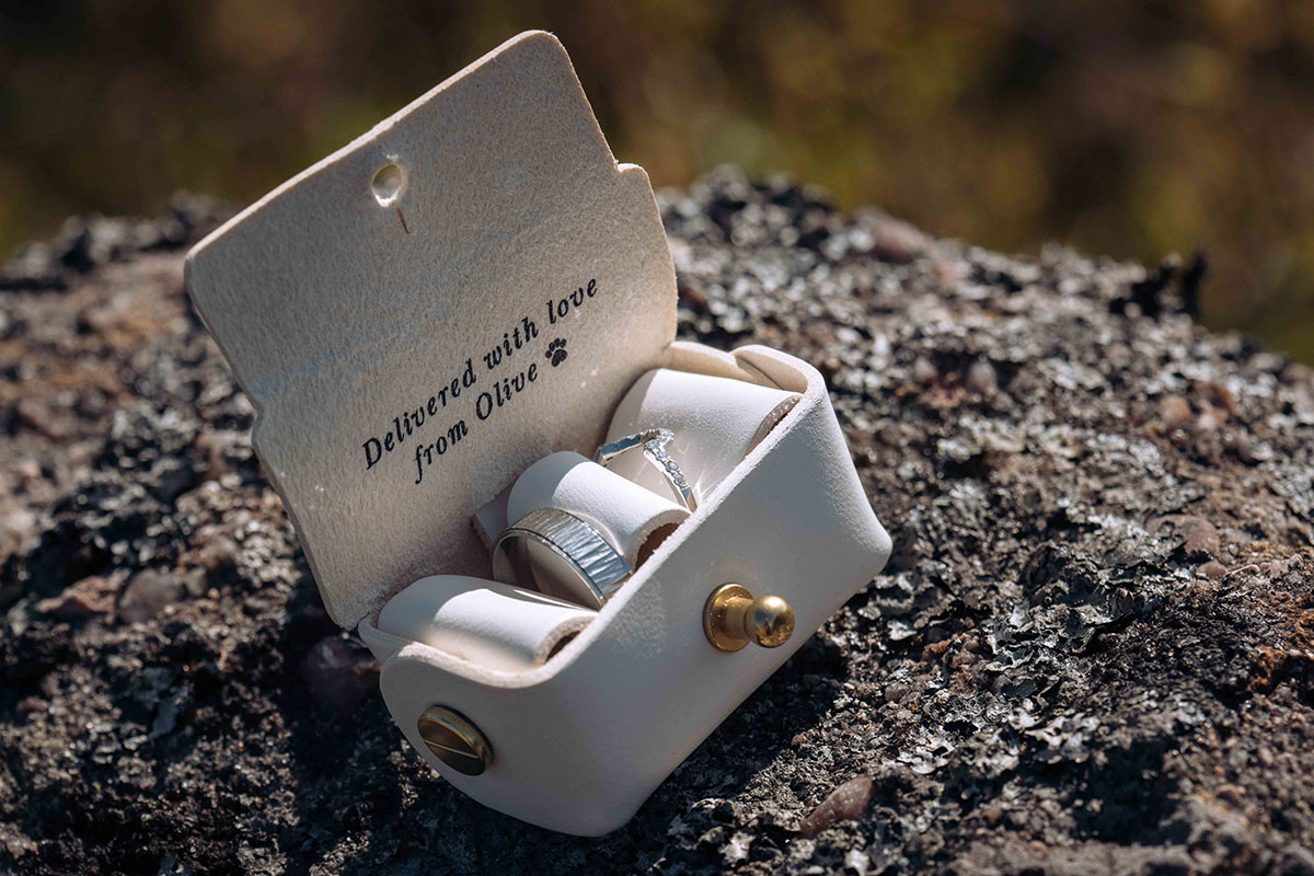 Close-up of the couple’s wedding rings in a cream leather box reading ‘Delivered with love from Olive’, their dog ring-bearer.