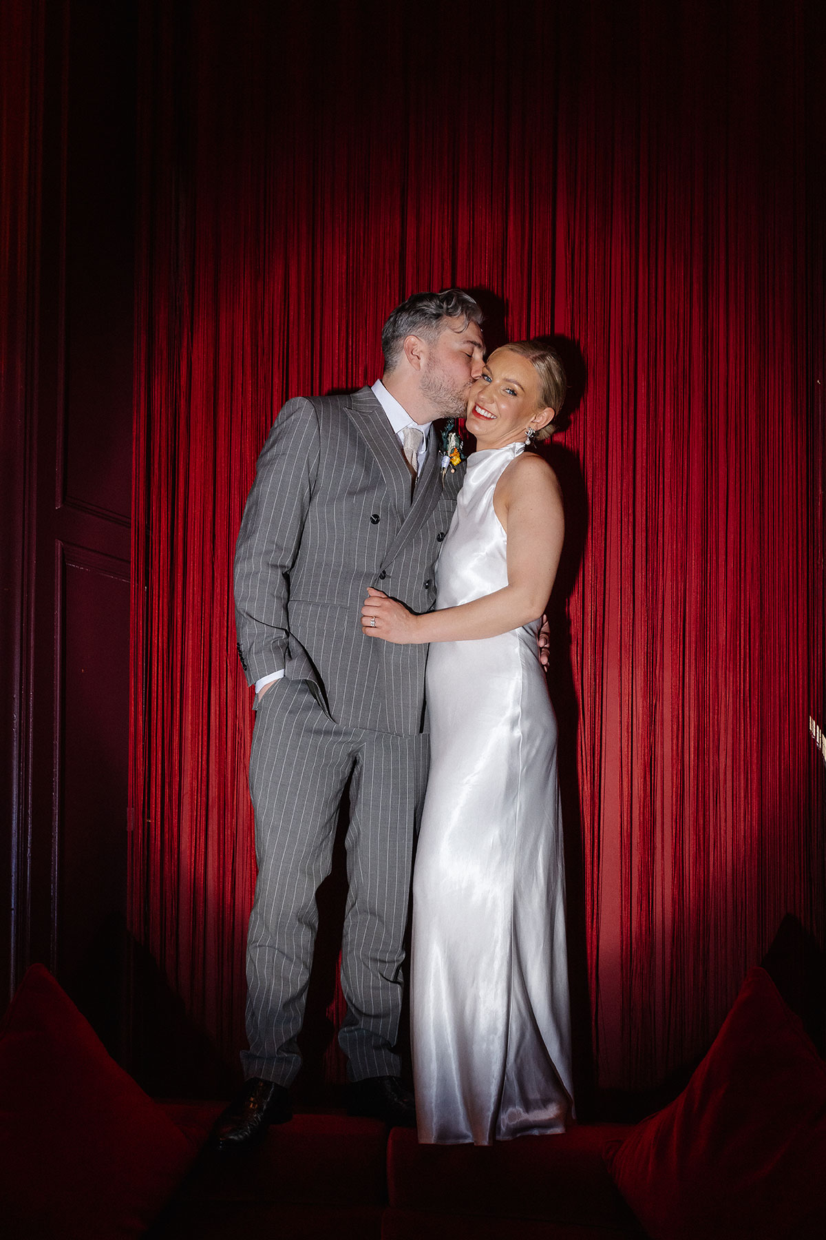 Bride and groom share a joyful moment in front of the red velvet backdrop inside the Scarlet Lounge at Virgin Hotels Edinburgh.