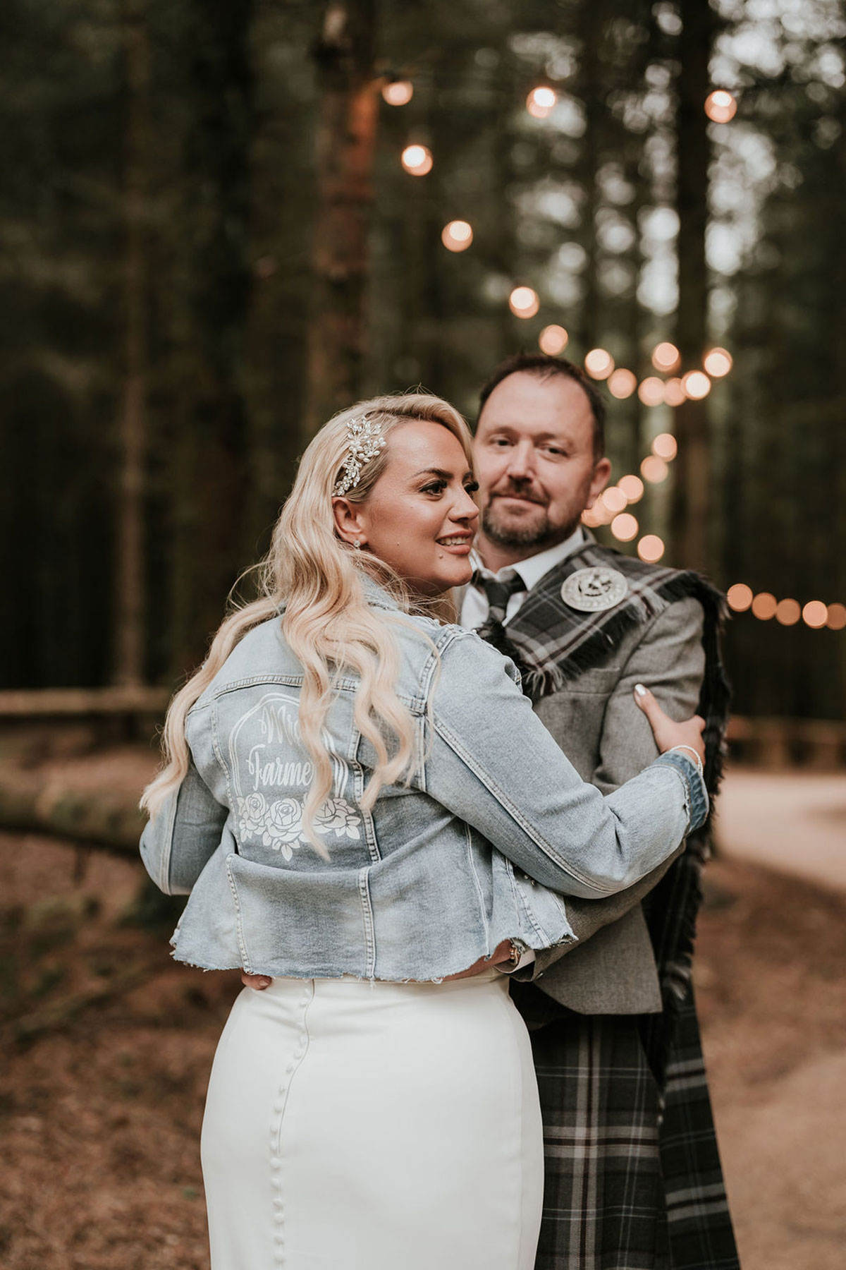 Bride wearing a personalised denim jacket while posing with the groom on a woodland path.