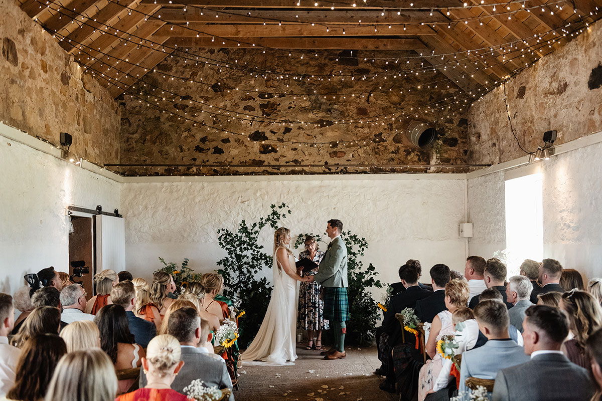 Bride and groom standing together at the altar with their celebrant, surrounded by guests seated in a fairy-lit barn